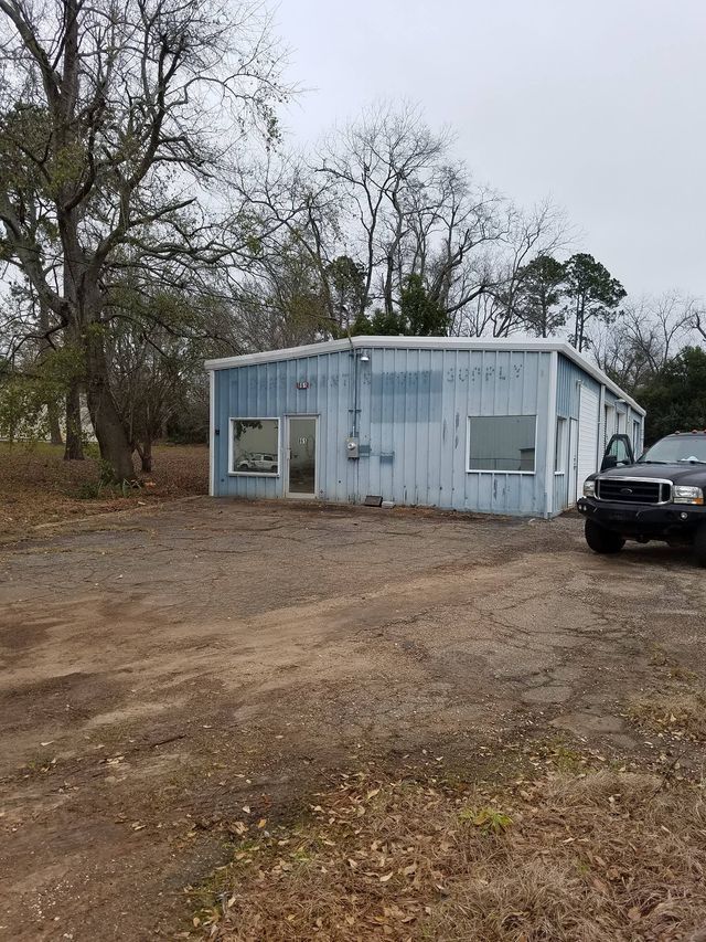 Small light-blue metal building in a dirt lot with trees around and part of a black truck visible on the right.