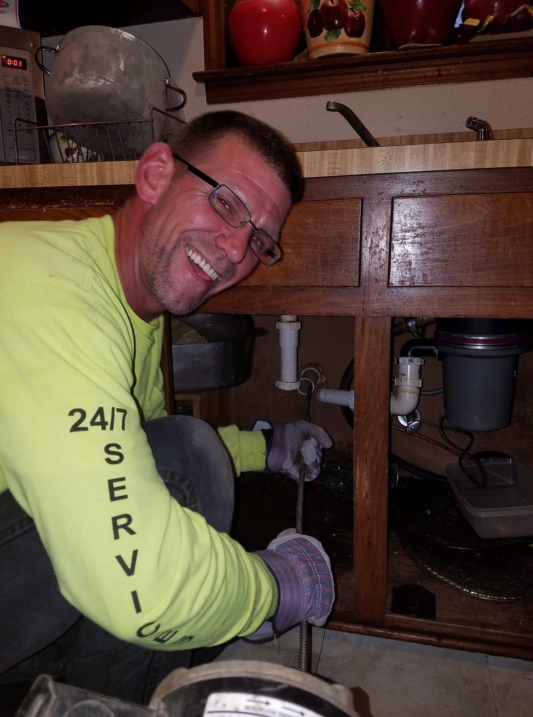 Man in neon yellow shirt kneels under a sink, smiling while working on plumbing with tools and purple gloves
