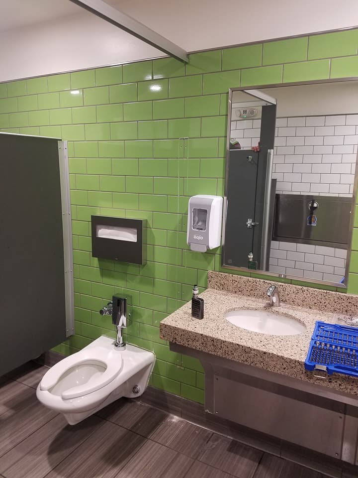 Public restroom with green tile wall, toilet, sink, mirror, and soap dispenser.