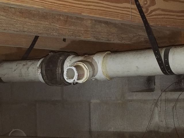 Exposed white PVC pipe with a repaired joint and clamp beneath wooden beams in a basement ceiling