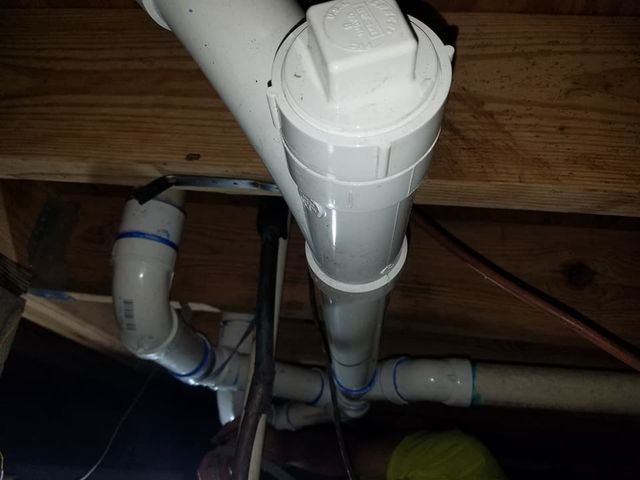 White PVC plumbing pipes and fittings under wooden floor joists in a crawl space.