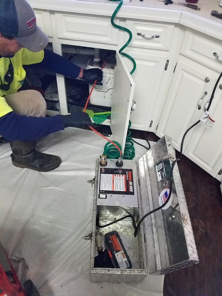 Technician working under a sink with tools and exposed wiring; a battery charger sits open on the floor.