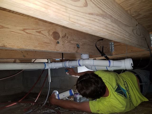 Technician working under a floor, installing white PVC plumbing pipes in a crawl space.