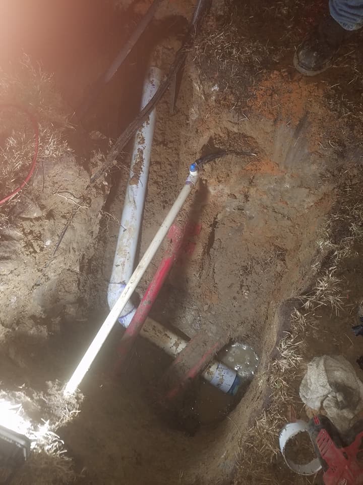 Trench with exposed underground pipes and cables in a muddy excavation site.