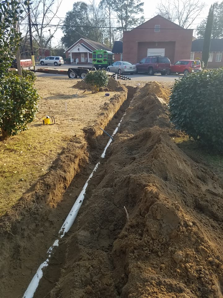 Trench dug through a yard toward a brick house, with exposed white pipe and dirt piled on both sides.