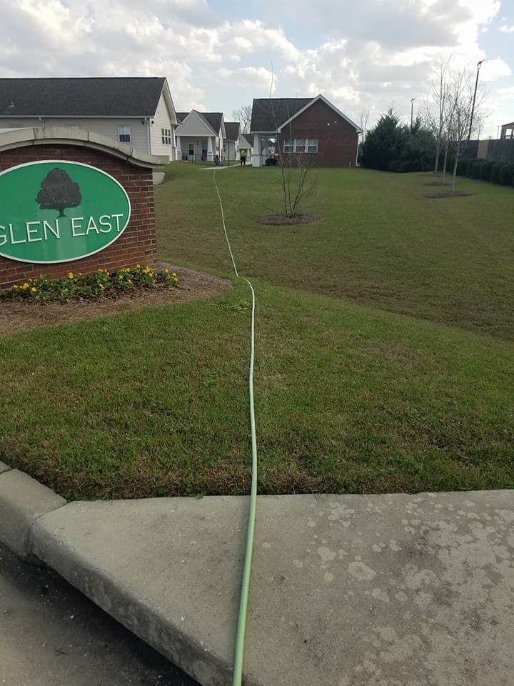 Green hose running across a grassy roadside entrance near the Glen Eys sign and houses.