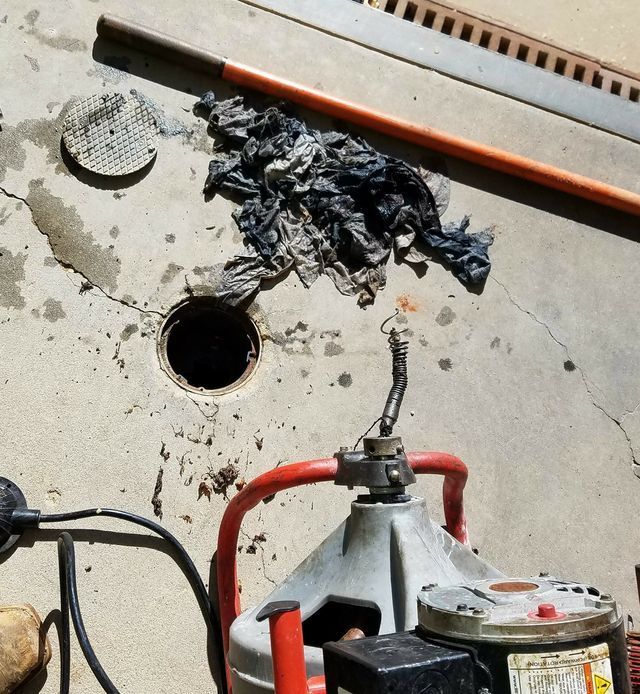 Open drain hole on concrete floor beside black debris, a red-handled tool, and a round metal cover.
