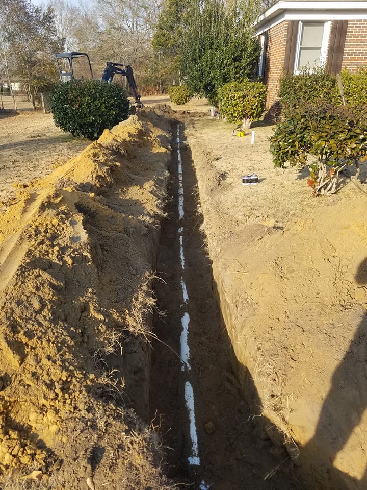 Trenched yard with a white pipe laid in the ground beside a house and shrubs