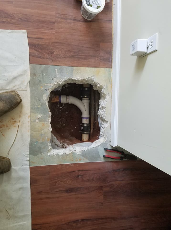 Floor cutout exposing plumbing beside a wall outlet in a room with wood flooring and a sofa edge