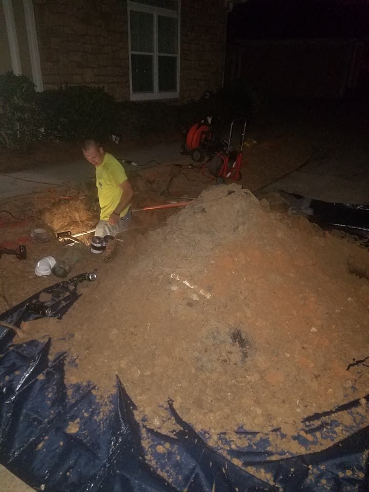 Nighttime construction site with a worker in a yellow jacket beside a large pile of dirt and tools on a tarp.