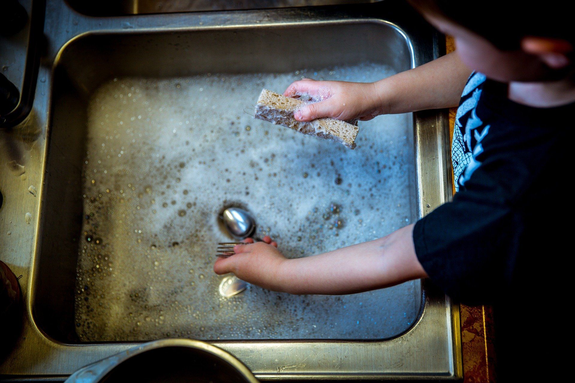 Hands washing a dish in a stainless steel kitchen sink with soap suds.