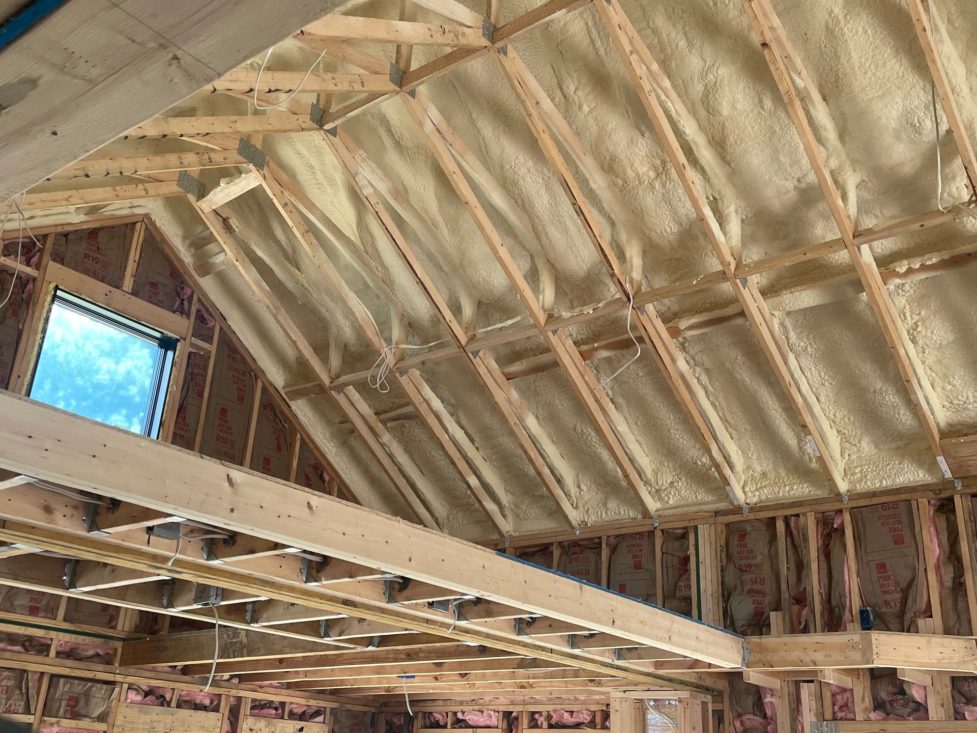 Exposed wooden roof framing and beams inside an unfinished building with spray insulation and a small window.