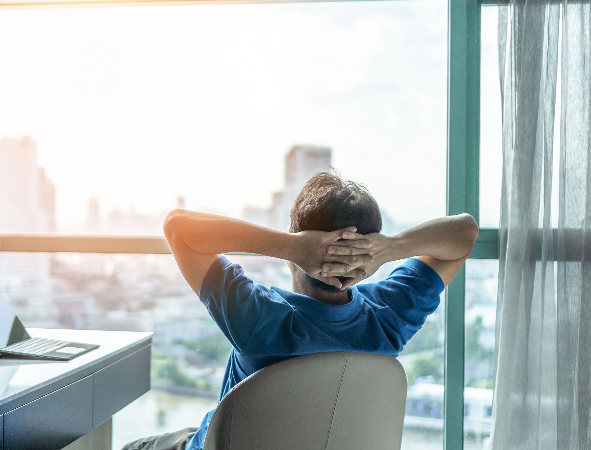 Person relaxing in chair by a window, arms behind head, overlooking a cityscape.