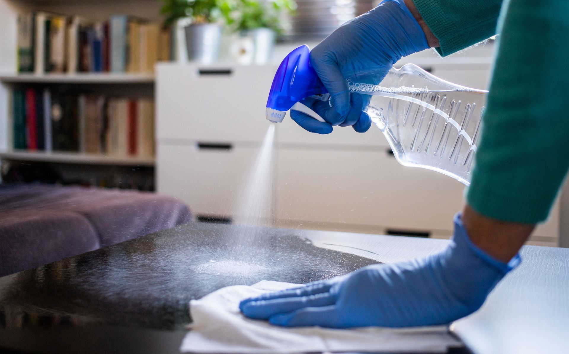 Hands in blue gloves spraying and wiping a dark surface with cleaning solution.
