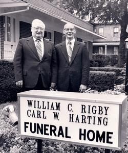 Two men stand beside a sign reading “William C. Rigby Carl W. Harting Funeral Home.”