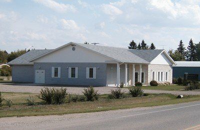 Single-story gray building with white columns and shrubs along a roadside on a sunny day
