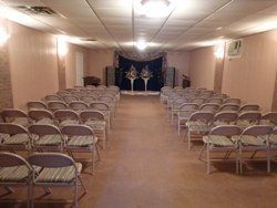 Empty banquet hall with rows of folding chairs facing a decorated stage under fluorescent lights