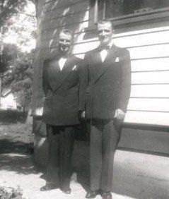 Two men in dark suits standing beside a white-sided house outdoors