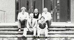 Four people seated on steps in front of a building entrance, posing for a black-and-white photo.