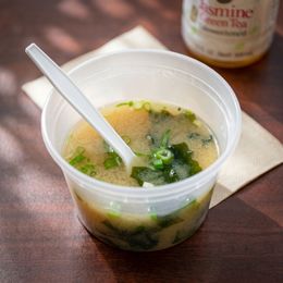 Plastic cup of soup with greens and a spoon on a wooden table, with a bottle blurred in the background