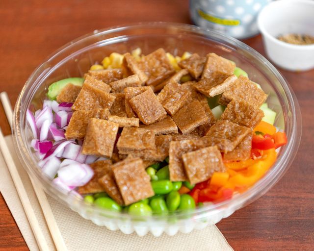 Colorful salad in a clear bowl with tofu cubes, red onion, peas, corn, and diced vegetables on a wooden table