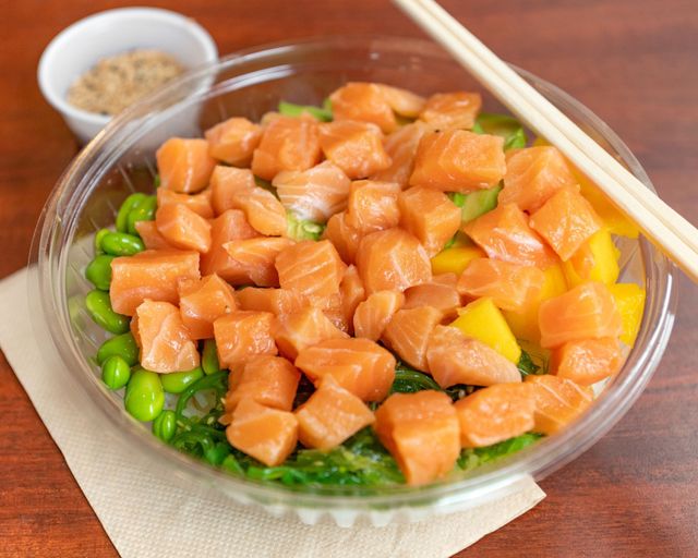 Salmon poke bowl with avocado, edamame, and rice in a clear takeaway container