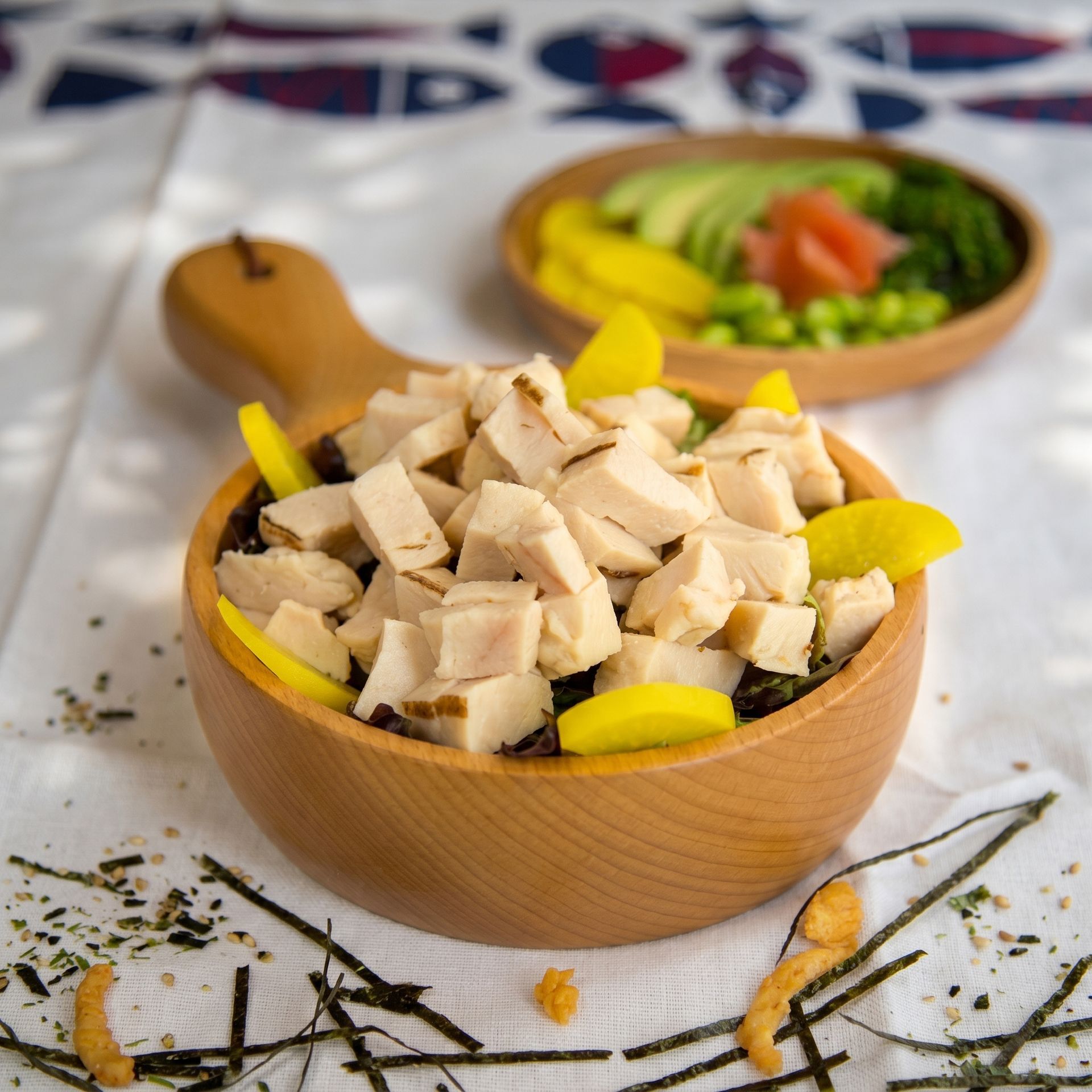 Wooden bowl of chopped chicken with lemon wedges on a white tablecloth, with a salad bowl in the background