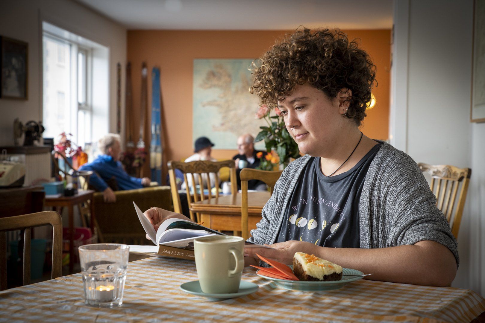 A woman is sitting at a table reading a book and eating a cake.