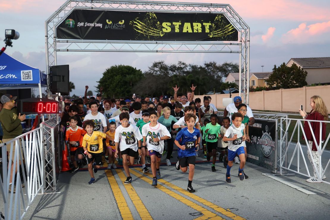 Runners of all ages starting a WildSide 5K race under a lit START arch at dusk, with RFID timing mats and finish-line equipment set up in the foreground