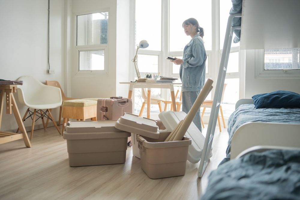 A person stands in a sunlit room filled with moving boxes and furniture, preparing for a move.