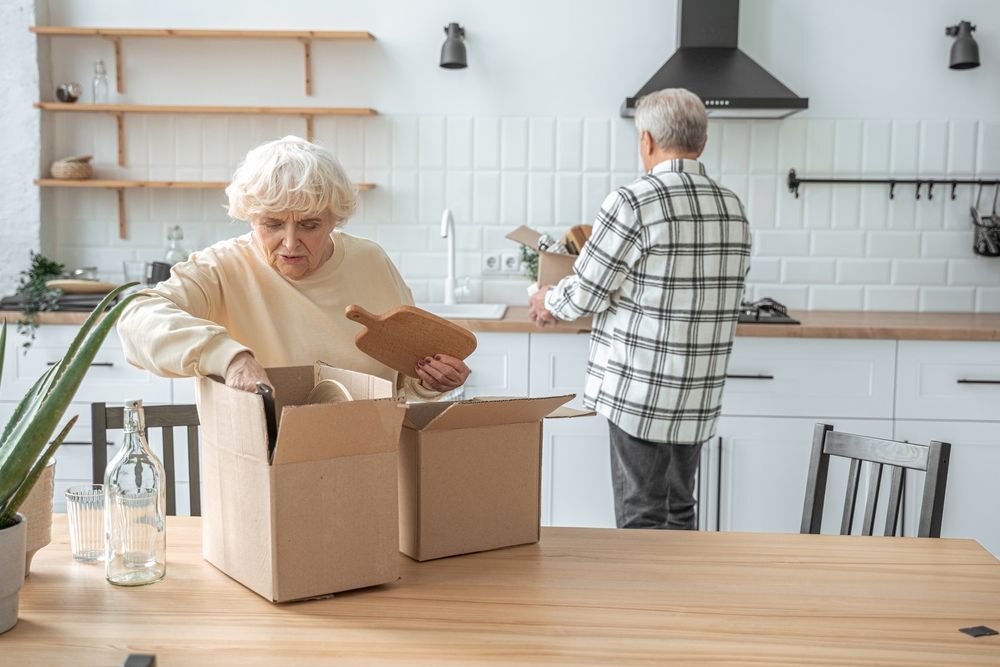 Two people pack kitchen items into cardboard boxes on a wooden table in a bright, modern kitchen.
