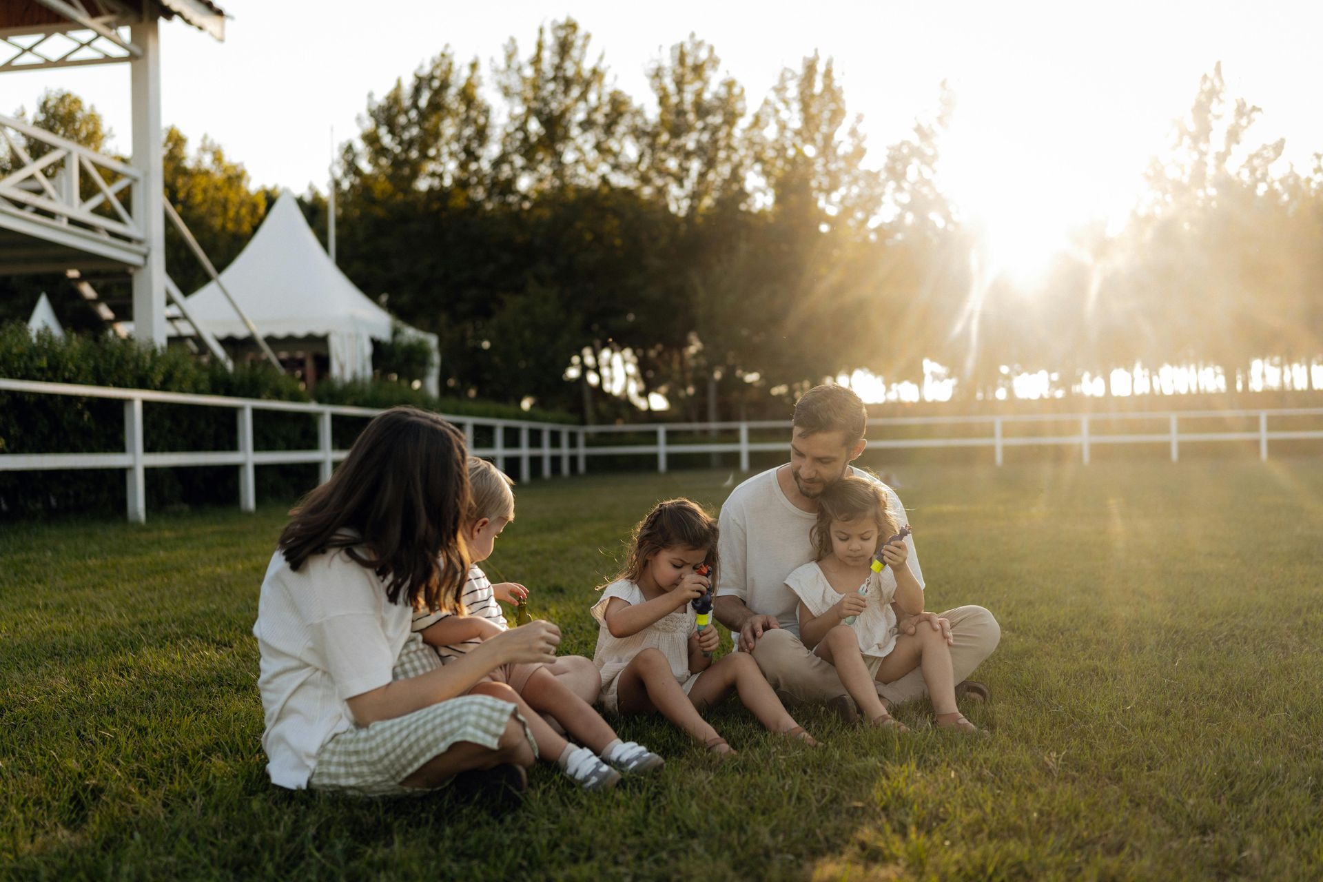 Family sitting on grass at sunset, sharing a picnic in a park near white fencing and trees.