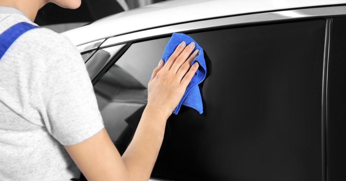 A person wiping a side car window with a cloth after applying tinted film inside an auto workshop bay.