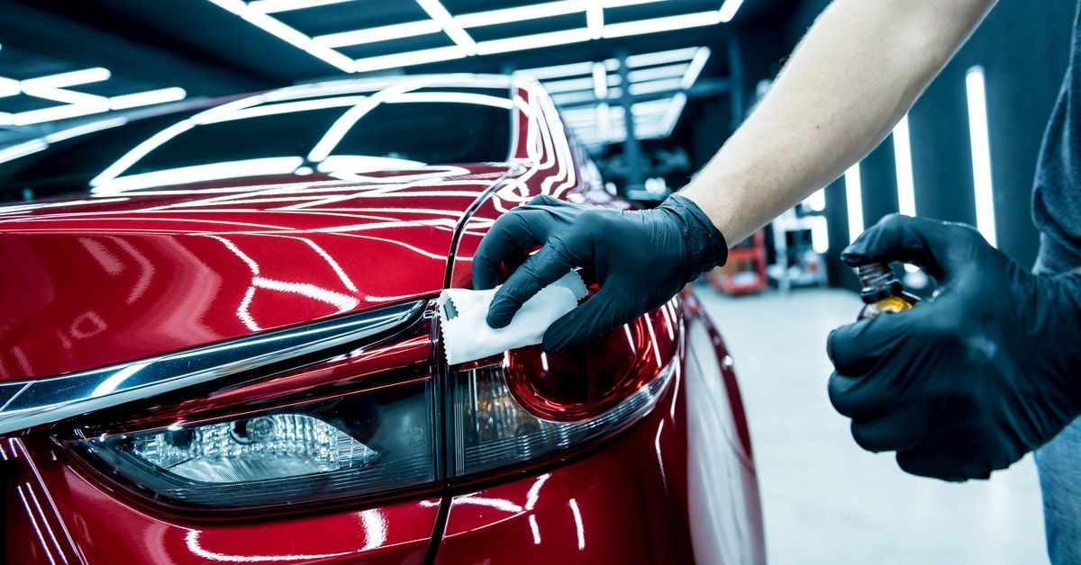 A car service worker wearing black gloves runs a ceramic coating along the front headlight of a red car.