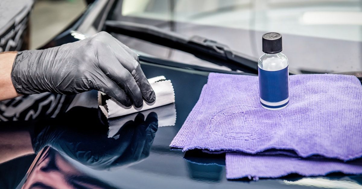 A specialist applies a ceramic coating over a scratch on a car. A bottle and a microfiber towel lay nearby.