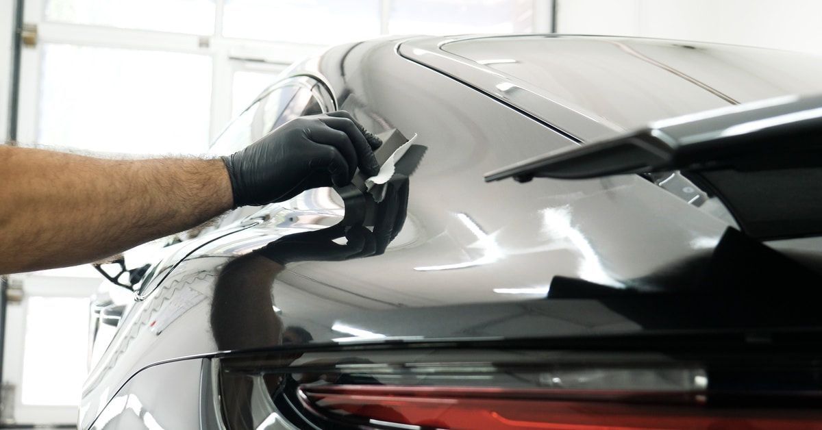 A person wearing black gloves applies a ceramic coating to a gray car. The vehicle is reflecting the light in the room.