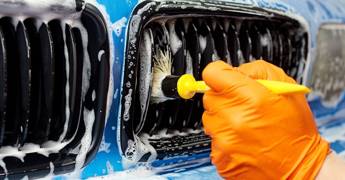 A hand with an orange glove on it holding a brush to clean the exterior of a car. The exterior is sudsy.