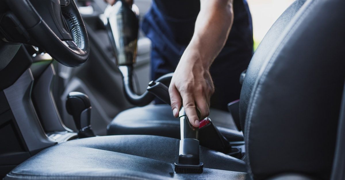 A person holding a vacuum and cleaning the driver's seat of a car. The vehicle's seats are black cloth.