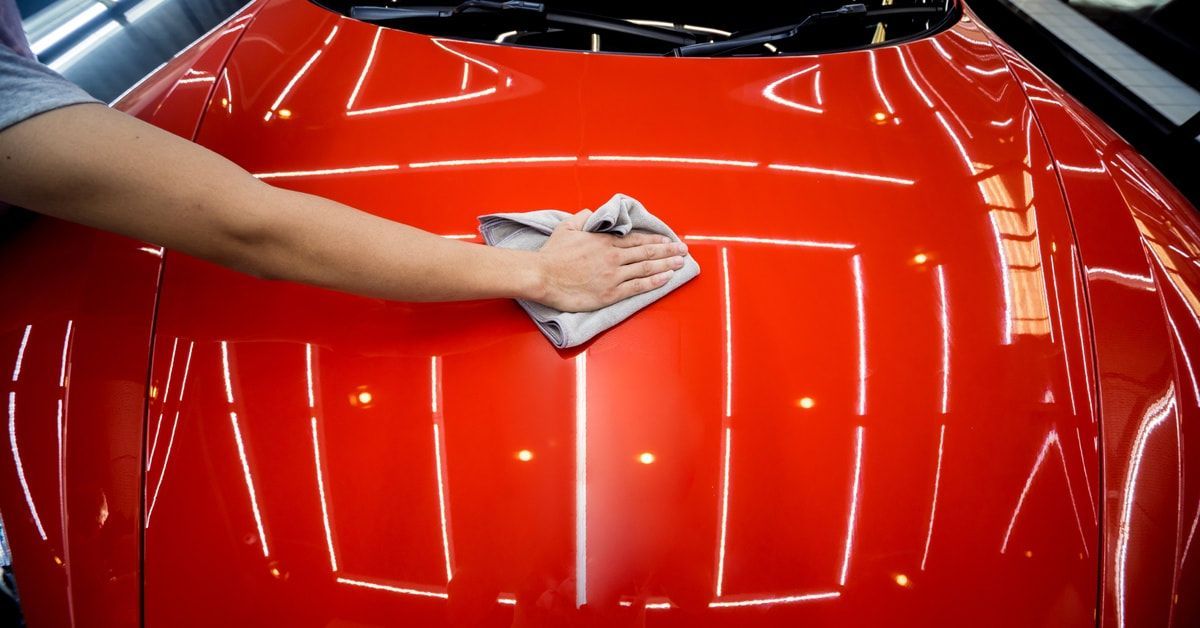 A person's hand running a microfiber towel over the hood of a red car. The car is so clean that it reflects the lights.