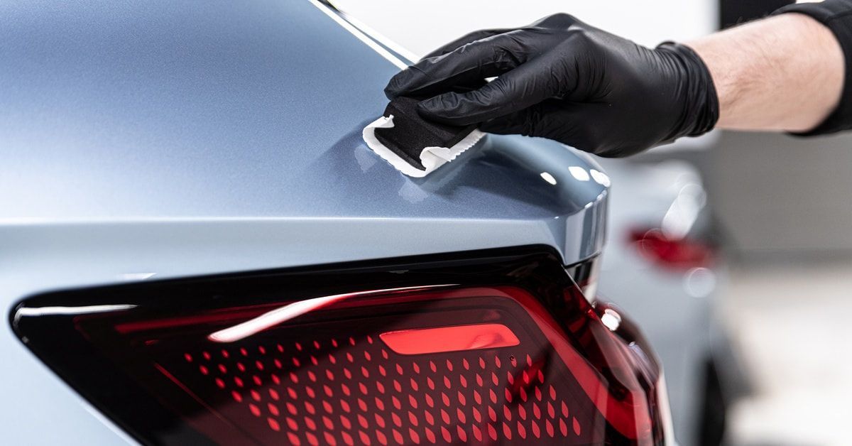 A car detailer applying a ceramic coating across the back of a light blue vehicle, above the taillight.