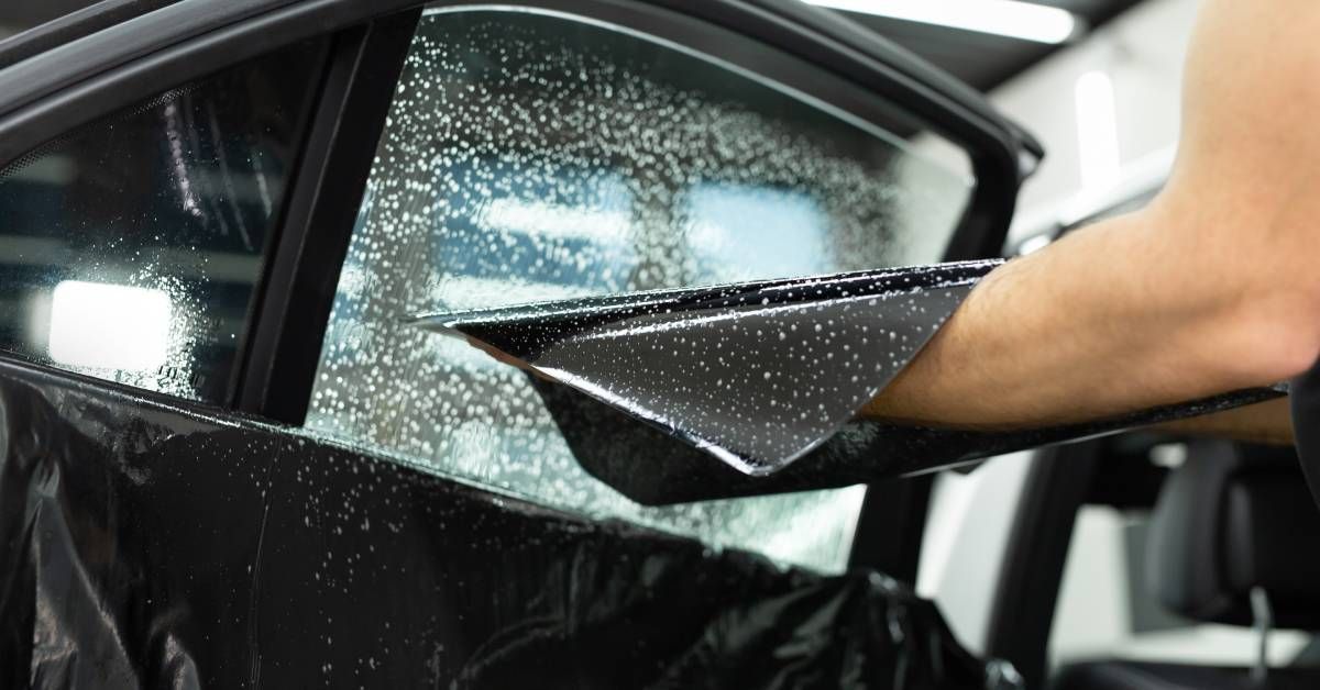 A vehicle window tinting service. The worker is holding a wet window tint next to a window covered in soap.