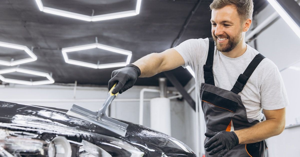 A smiling man washing the hood of a car and preparing it for protective film. He is wearing gloves and an apron.