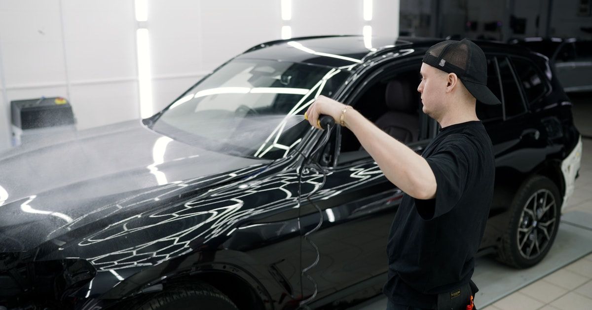 Professional auto detailer spraying a glossy black SUV with a hose. He is wearing all black and in a workshop.