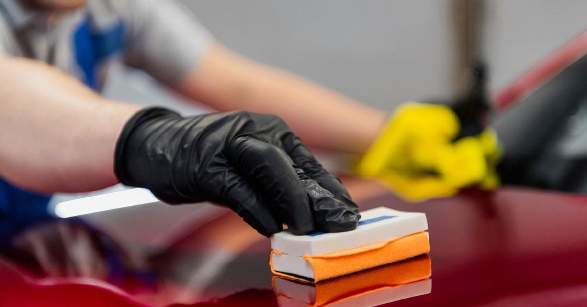 A man wearing black gloves is running a sponge along a red car to apply the protective ceramic coating.