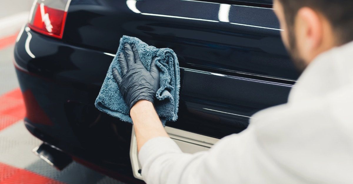 A person wiping the rear of a black car with a cloth inside an auto service bay with bright overhead lights.