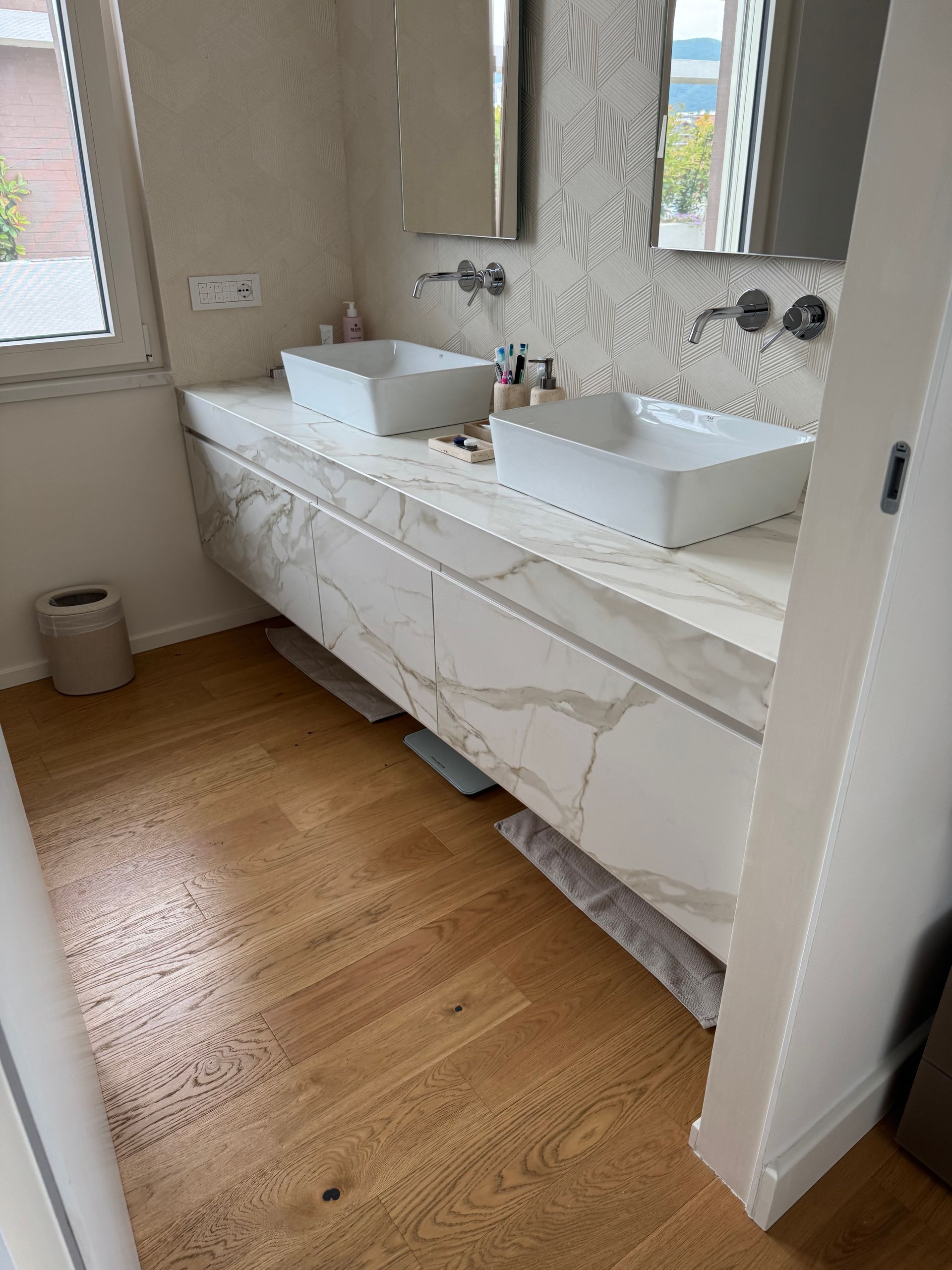 Bagno moderno con doppio lavabo, ampio specchio, mobiletto in marmo e pavimento effetto legno.