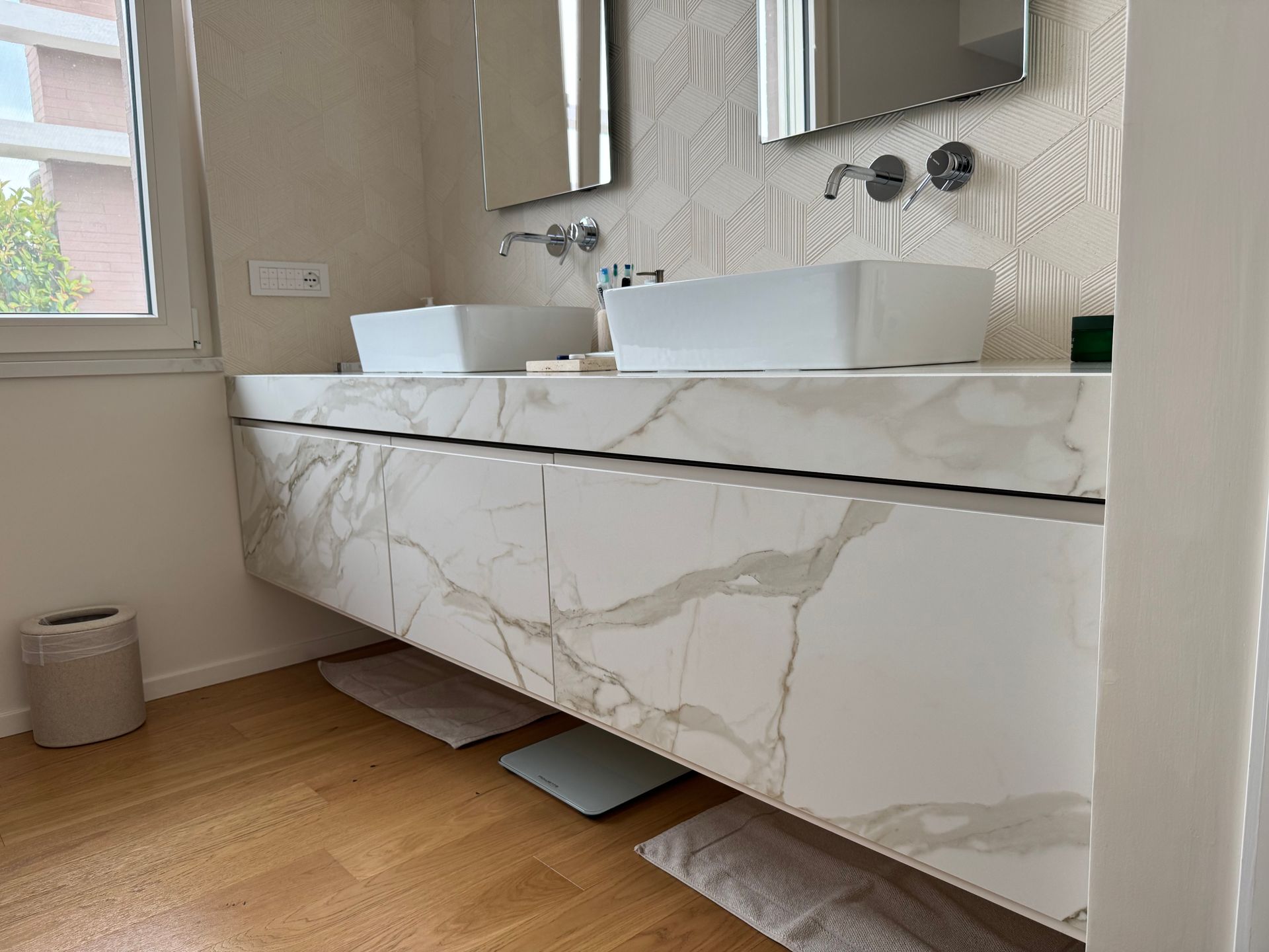 Mobile bagno moderno con cassetti effetto marmo, doppio lavabo e finestra sopra il pavimento in legno.