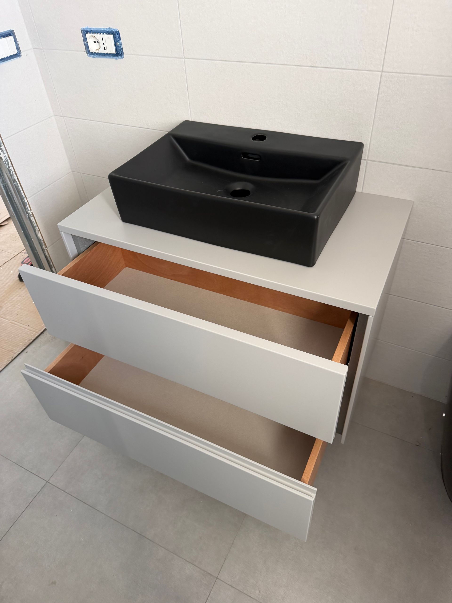 Mobile da bagno con lavabo nero e cassetti bianchi a giorno, che mostrano l'interno in legno non trattato.