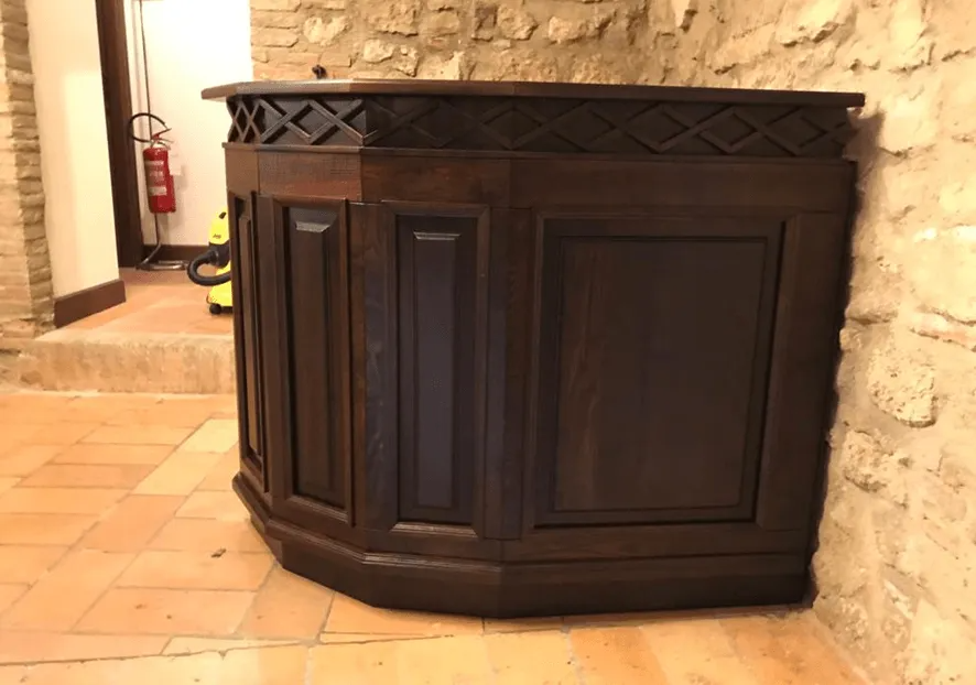 Bancone bar in legno scuro con finiture decorative, addossato a una parete in pietra in un ambiente interno rustico.