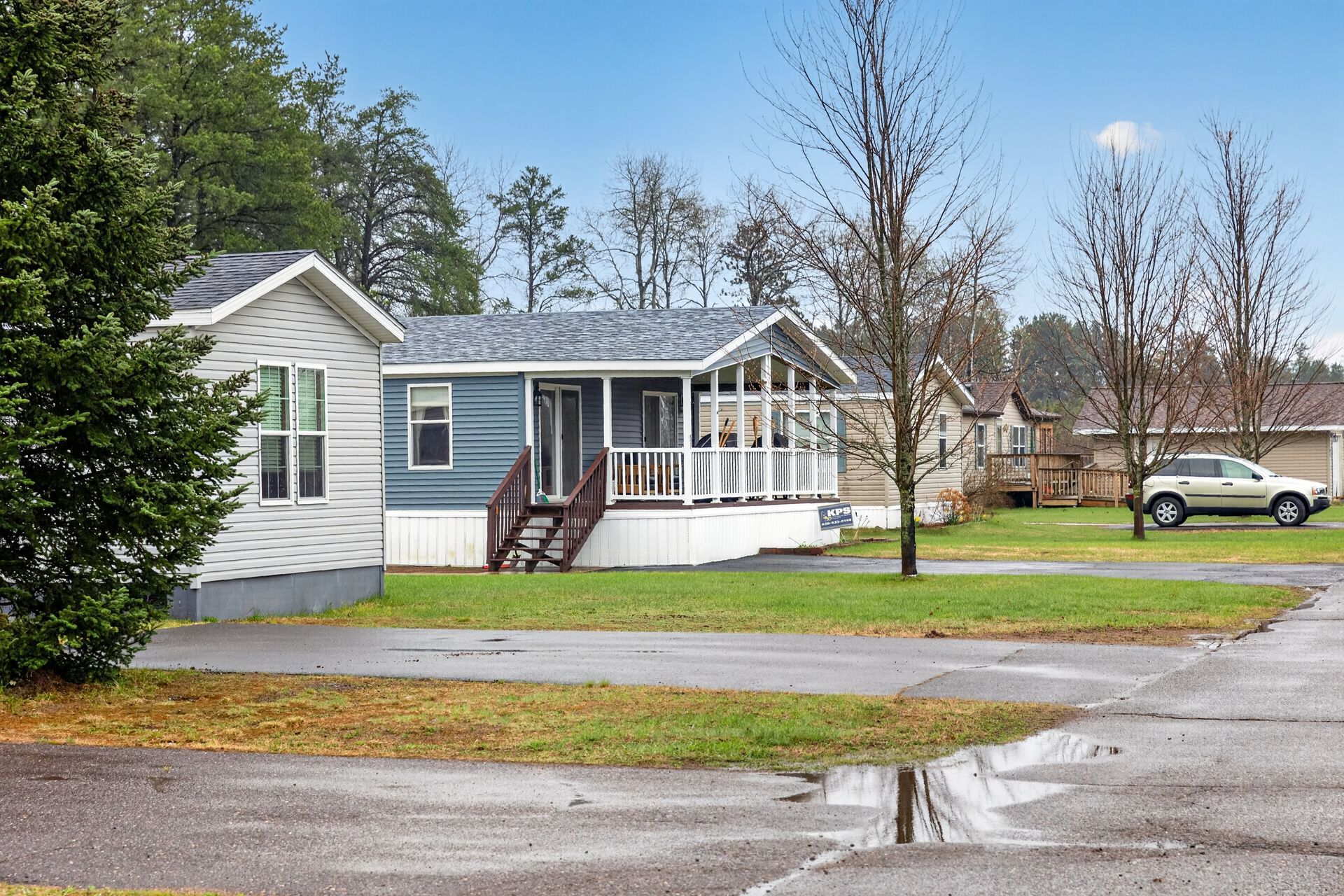 Small white house with blue siding and front porch beside a wet street and parked car.